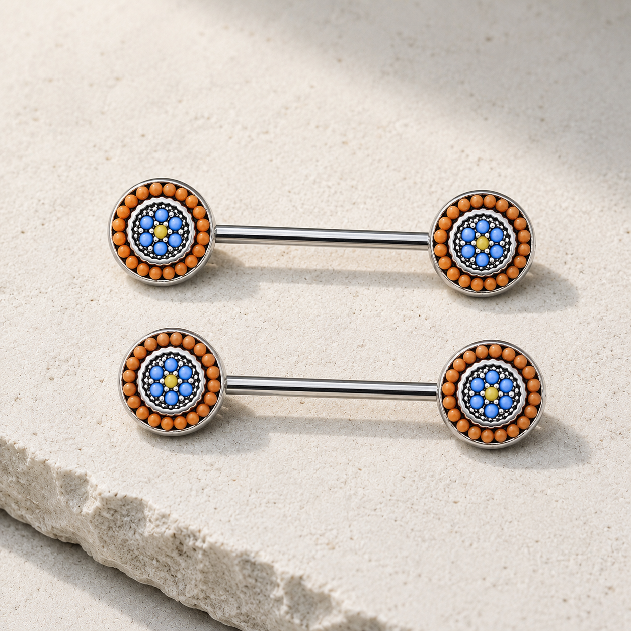 Two metallic nipple barbells with blue and orange bead designs on a stone surface.