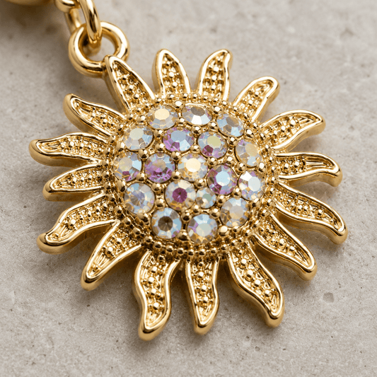 A gold sunburst pendant charm with a central cluster of iridescent round rhinestones, shown close-up on a textured light gray surface.
